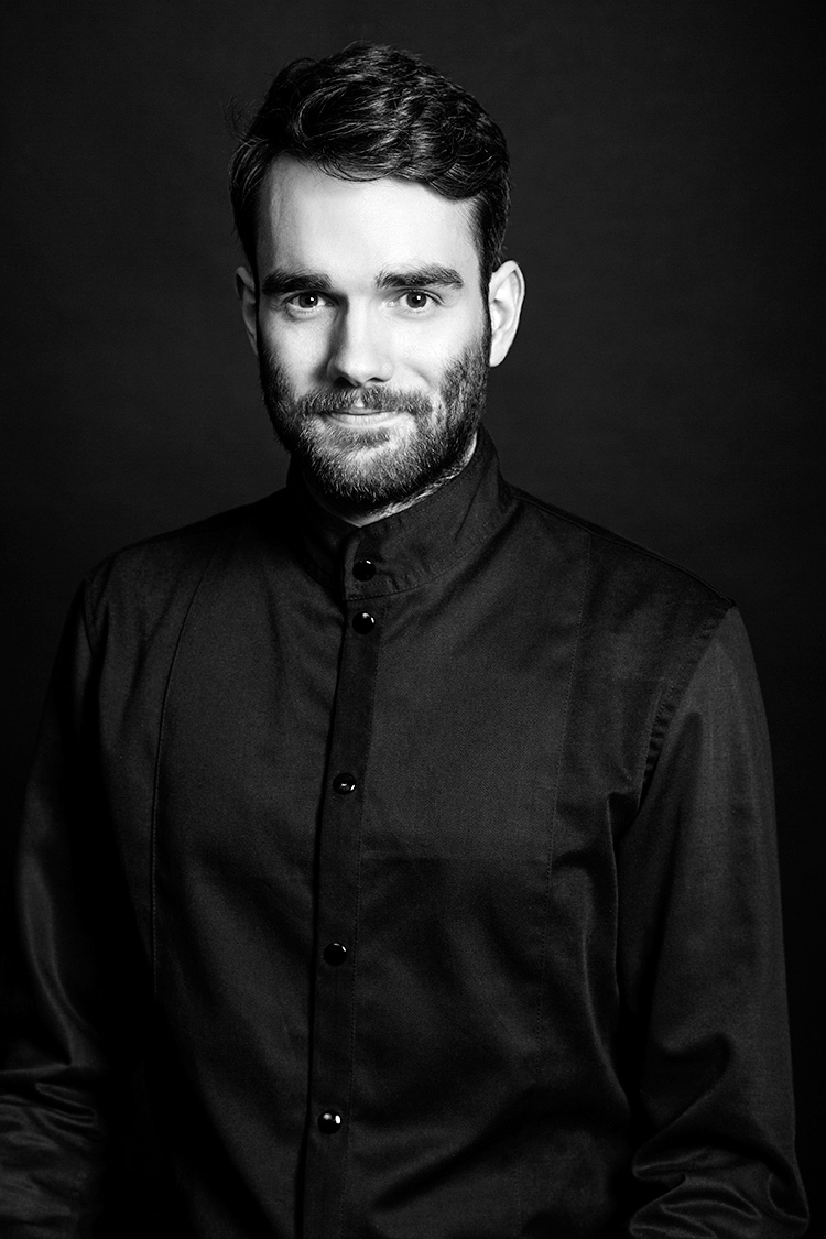 Filip Jakobsen, CEO & founder. Black and white photo with contrastful lighting and a dark grey background. Shows a man in his late twenties with light skin, a serious expression and slight smile. The man has short, dark hair and a short full beard, is wearing a black standing collar shirt, and is looking into the camera.