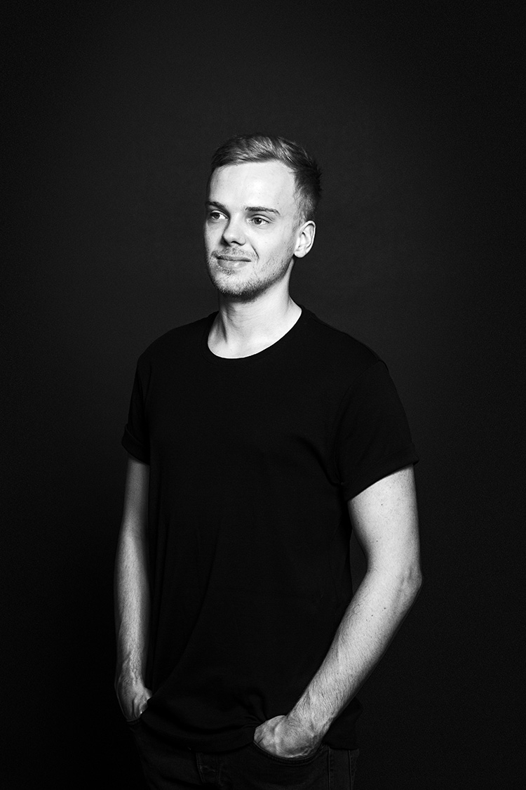Rasmus Wølk, UX developer. Black and white photo with contrastful lighting and a dark grey background. Shows a young man in his late twenties with light skin and short, dark blond hair, wearing a black t-shirt. The man has a slight smile, is facing the camera with his face and eyes pointed slightly left.